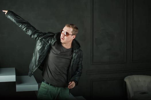 studio photography young brutal guy. man in sunglasses, T-shirt, jeans puts on black leather jacket. black background painted walls and white stairs