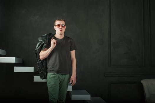 studio photography young brutal guy. man in sunglasses, T-shirt, jeans keeps black leather jacket, slung over his shoulder. black background painted walls and white stairs