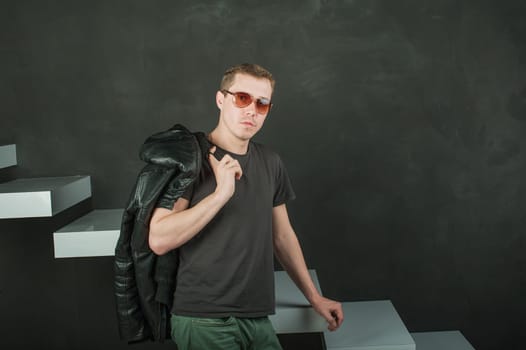 studio photography young brutal guy. man in sunglasses, T-shirt, jeans keeps black leather jacket, slung over his shoulder. black background painted walls and white stairs