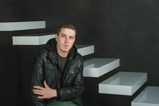 studio photography of a young man in a dark T-shirt, black leather jacket. man sitting looking at the camera with arms crossed against a background of dark walls and white stairs