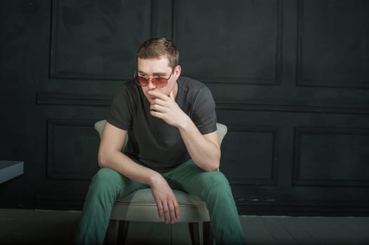 studio photography young brutal guy. man in sunglasses, T-shirt, jeans and high boots on a background of black wall, He is sitting in a white armchair throne.