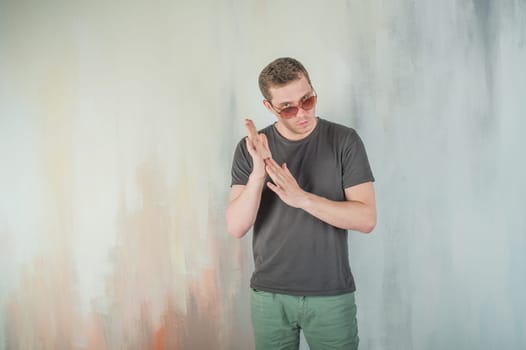 studio photography young brutal guy. man in sunglasses, T-shirt, jeans on a background colorful painted walls. his hands clasped palm to palm rubbing them