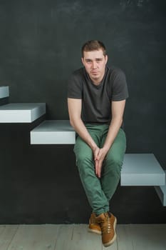 studio photography young brutal guy. man in T-shirt, jeans and high boots on a background of black wall on the white steps. sitting on the steps, his hands clasped palm to palm, looking at the camera