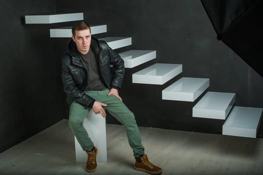 studio photography of a young man in a dark T-shirt, black leather jacket. man sitting, made of gun fingers against the dark walls and white stairs
