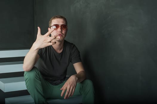 studio photography young brutal guy. man in sunglasses, T-shirt, jeans and high boots on a background of black wall on the white steps. sitting on the steps, shows hands