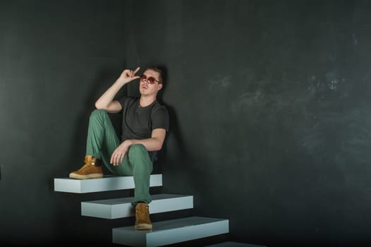 studio photography young brutal guy. man in sunglasses, T-shirt, jeans and high boots on a background of black wall on the white steps. sitting on the steps