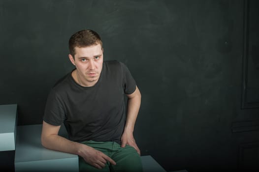 studio photography young brutal guy. man in T-shirt on a background of black wall on the white steps. sitting on the steps, looking at the camera