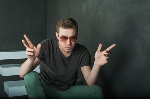 studio photography young brutal guy. man in sunglasses, T-shirt, jeans and high boots on a background of black wall on the white steps. sitting on the steps, shows hands