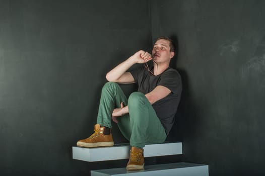 studio photography young brutal guy. man in sunglasses, T-shirt, jeans and high boots on a background of black wall on the white steps. sitting on the steps, took off his glasses and holds them in his hand
