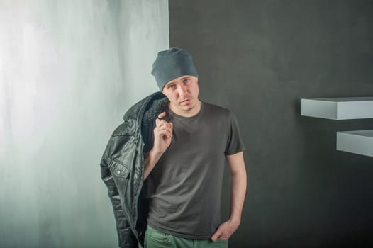 studio photography young brutal guy. man in T-shirt, jeans and a knitted cap, keeps black leather jacket, slung over his shoulder. black background painted walls and white stairs