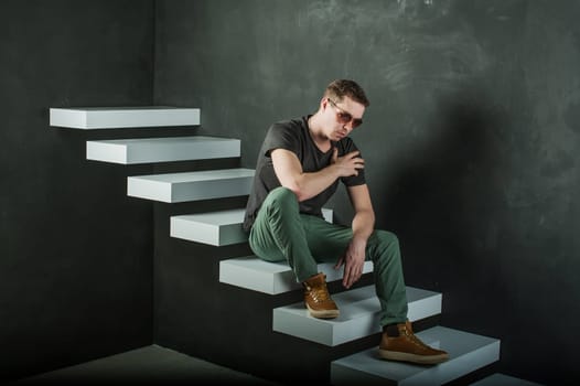 studio photography young brutal guy. man in sunglasses, T-shirt, jeans and high boots on a background of black wall on the white steps. sitting on the stairs, looked down, lightly touching his shoulder with his hand