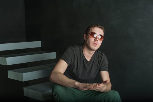 studio photography young brutal guy. man in sunglasses, T-shirt, jeans and high boots on a background of black wall on the white steps. sitting on the steps, his hands clasped palm to palm rubbing them