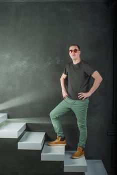 studio shooting young brutal guy. man in sunglasses, T-shirt, jeans and high boots on a background of black wall on the white steps. He is looking into the camera, standing arms akimbo