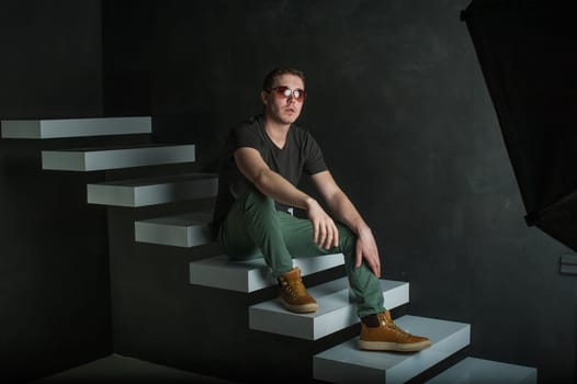 studio photography young brutal guy. man in sunglasses, T-shirt, jeans and high boots on a background of black wall on the white steps. sitting on the steps, protinuv hands forward, putting them on her knees
