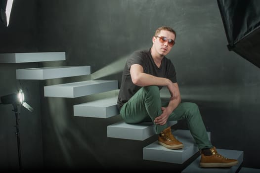 studio photography young brutal guy. man in sunglasses, T-shirt, jeans and high boots on a background of black wall on the white steps. He is looking at the camera while sitting on the stairs, hand on knee