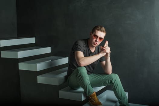 studio photography young brutal guy. man in sunglasses, T-shirt, jeans and high boots on a background of black wall on the white steps. sitting on the steps, holding hands in the shape of a gun for a shot
