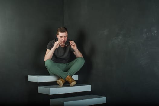 studio photography young brutal guy. man in sunglasses, T-shirt, jeans and high boots on a background of black wall on the white steps. sitting on the steps, took off his glasses and holds them in his hand