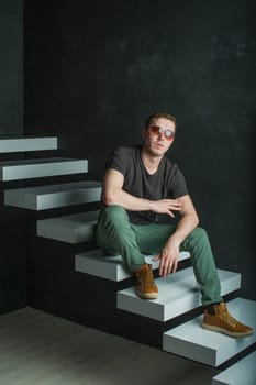 studio photography young brutal guy. man in sunglasses, T-shirt, jeans and high boots on a background of black wall on the white steps. He is looking at the camera while sitting on the stairs, hand on knee