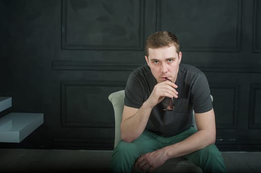 studio photography young brutal guy. man in sunglasses, T-shirt, jeans and high boots on a background of black wall, He is sitting in a white armchair throne.