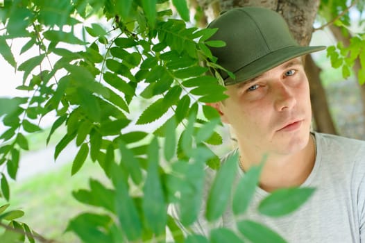 young guy standard physique posing on the street on a background of green bushes. He is wearing a light shirt and dark green cap. portrait of a man summer day