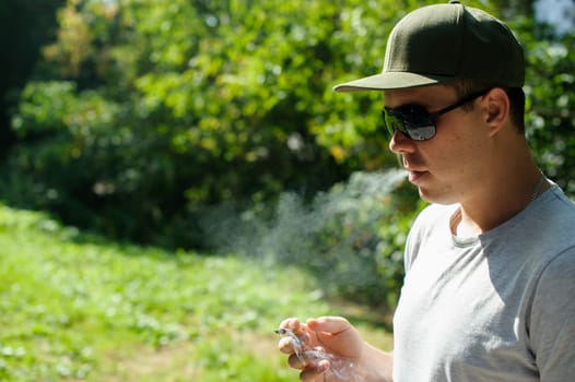 a young man dressed in light and dark ftbolku cap, smoking a cigarette emitting smoke in the background of summer green sunny day