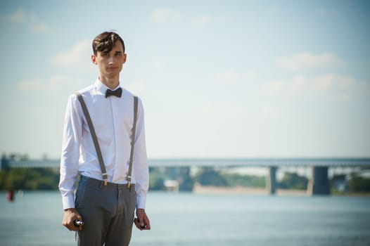 young skinny man, elegantly stylishly dressed in white shirt, trousers with suspenders and bow tie. vaper guy Smokes an e-cigarette, releas vapors of smoke, against background of river and bridge