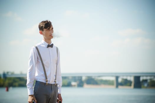 young skinny man, elegantly stylishly dressed in white shirt, trousers with suspenders and bow tie. vaper guy Smokes an e-cigarette, releas vapors of smoke, against background of river and bridge