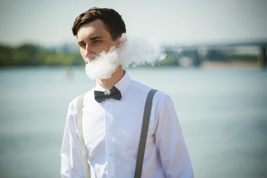 young skinny man, elegantly stylishly dressed in white shirt, trousers with suspenders and bow tie. vaper guy Smokes an e-cigarette, releas vapors of smoke, against background of river and bridge