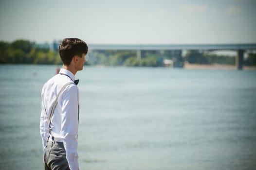 young skinny man, elegantly stylishly dressed in white shirt, trousers with suspenders and bow tie. vaper guy Smokes an e-cigarette, releas vapors of smoke, against background of river and bridge