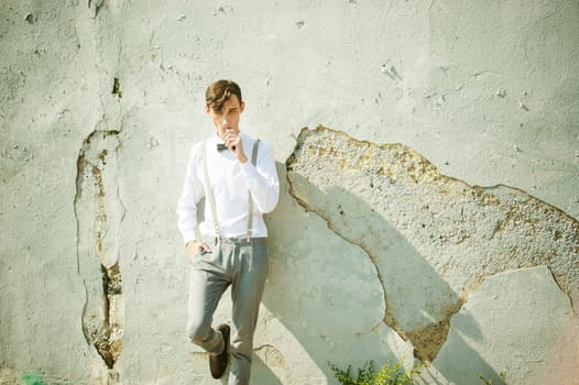young skinny man, elegantly stylishly dressed in white shirt, gray trousers with suspenders and bow tie. Smokes an electronic cigarette on background of shabby wall, warm, sunny summer day