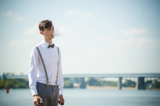 young skinny man, elegantly stylishly dressed in white shirt, trousers with suspenders and bow tie. vaper guy Smokes an e-cigarette, releas vapors of smoke, against background of river and bridge