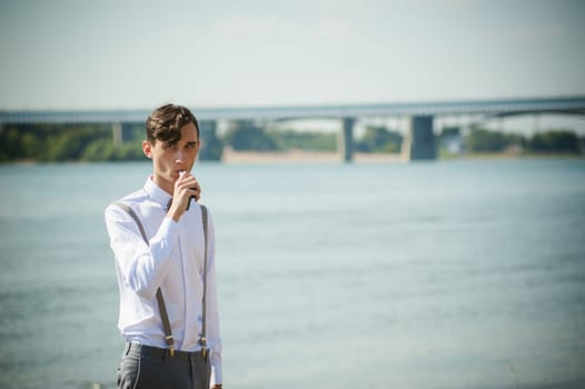 young skinny man, elegantly stylishly dressed in white shirt, trousers with suspenders and bow tie. vaper guy Smokes an e-cigarette, releas vapors of smoke, against background of river and bridge