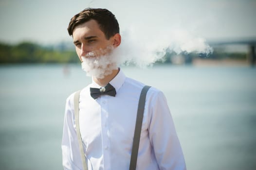 young skinny man, elegantly stylishly dressed in white shirt, trousers with suspenders and bow tie. vaper guy Smokes an e-cigarette, releas vapors of smoke, against background of river and bridge