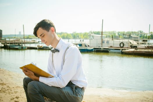 young skinny man, elegantly stylishly dressed in white shirt, gray trousers with suspenders and bow tie. young guy on pier on background of river and boats, takes notes of pen on sheet in notebook