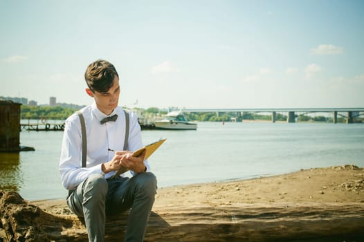 young skinny man, elegantly stylishly dressed in white shirt, gray trousers with suspenders and bow tie. young guy on pier on background of river and boats, takes notes of pen on sheet in notebook