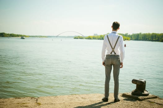 young skinny man, elegantly stylishly dressed in white shirt, gray trousers with suspenders and bow tie. Looking away, at the river water bridge and ship