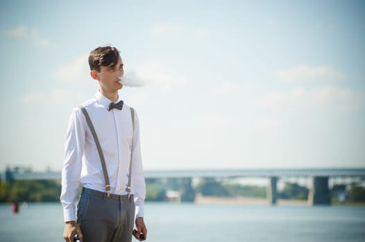 young skinny man, elegantly stylishly dressed in white shirt, trousers with suspenders and bow tie. vaper guy Smokes an e-cigarette, releas vapors of smoke, against background of river and bridge