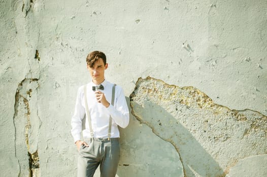 young skinny man, elegantly stylishly dressed in white shirt, gray trousers with suspenders and bow tie. Smokes an electronic cigarette on background of shabby wall, warm, sunny summer day