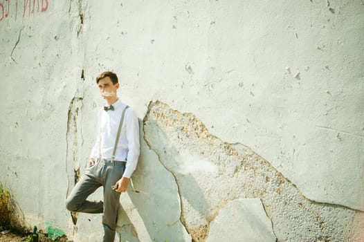 young skinny man, elegantly stylishly dressed in white shirt, gray trousers with suspenders and bow tie. Smokes an electronic cigarette on background of shabby wall, warm, sunny summer day