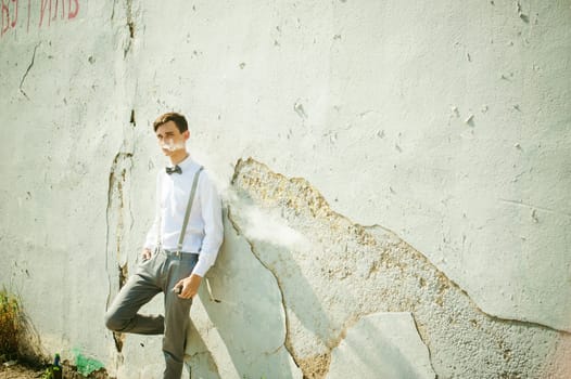 young skinny man, elegantly stylishly dressed in white shirt, gray trousers with suspenders and bow tie. Smokes an electronic cigarette on background of shabby wall, warm, sunny summer day