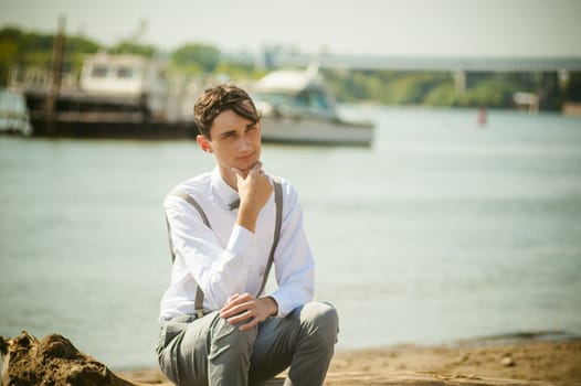 young skinny man, elegantly stylishly dressed in white shirt, gray trousers with suspenders and bow tie. Portrait young guy on pier on background of river and boats