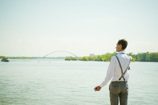 young skinny man, elegantly stylishly dressed in white shirt, gray trousers with suspenders and bow tie. Looking away, at the river water bridge and ship