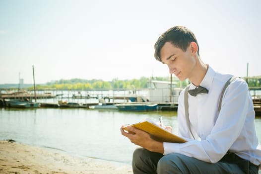 young skinny man, elegantly stylishly dressed in white shirt, gray trousers with suspenders and bow tie. young guy on pier on background of river and boats, takes notes of pen on sheet in notebook