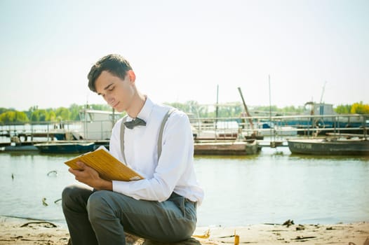 young skinny man, elegantly stylishly dressed in white shirt, gray trousers with suspenders and bow tie. young guy on pier on background of river and boats, takes notes of pen on sheet in notebook