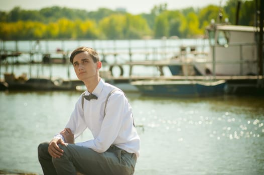 young skinny man, elegantly stylishly dressed in white shirt, gray trousers with suspenders and bow tie. Portrait young guy on pier on background of river and boats