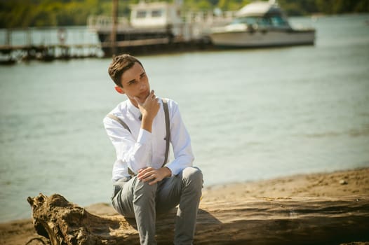 young skinny man, elegantly stylishly dressed in white shirt, gray trousers with suspenders and bow tie. Portrait young guy on pier on background of river and boats