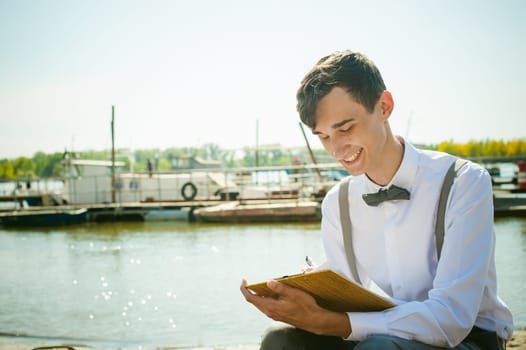 young skinny man, elegantly stylishly dressed in white shirt, gray trousers with suspenders and bow tie. young guy on pier on background of river and boats, takes notes of pen on sheet in notebook