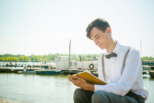 young skinny man, elegantly stylishly dressed in white shirt, gray trousers with suspenders and bow tie. young guy on pier on background of river and boats, takes notes of pen on sheet in notebook
