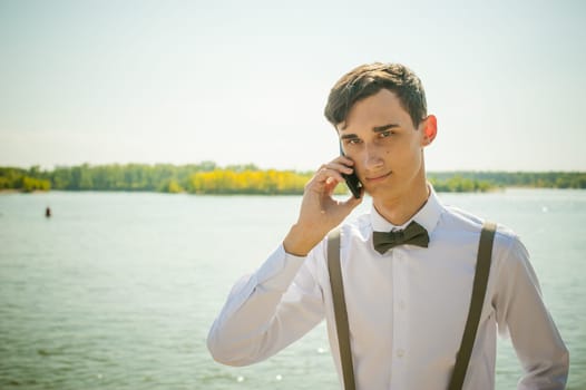 young skinny man, elegantly stylishly dressed in white shirt, gray trousers with suspenders and bow tie. Portrait of young guy talking on phone on background of river on warm sunny summer day
