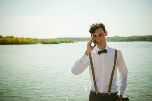 young skinny man, elegantly stylishly dressed in white shirt, gray trousers with suspenders and bow tie. Portrait of young guy talking on phone on background of river on warm sunny summer day